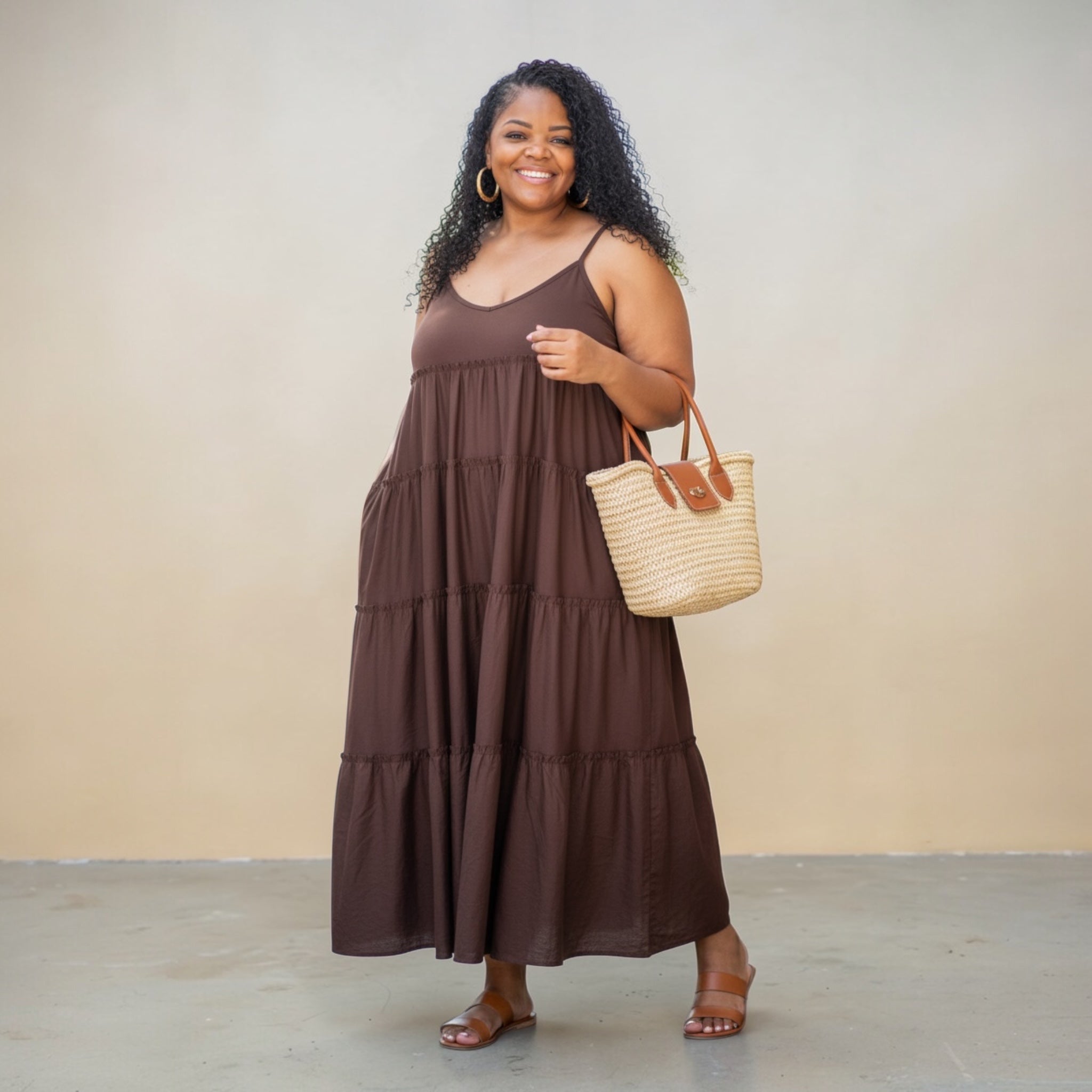 Woman wearing a brown tiered dress holding a straw bag against a beige background